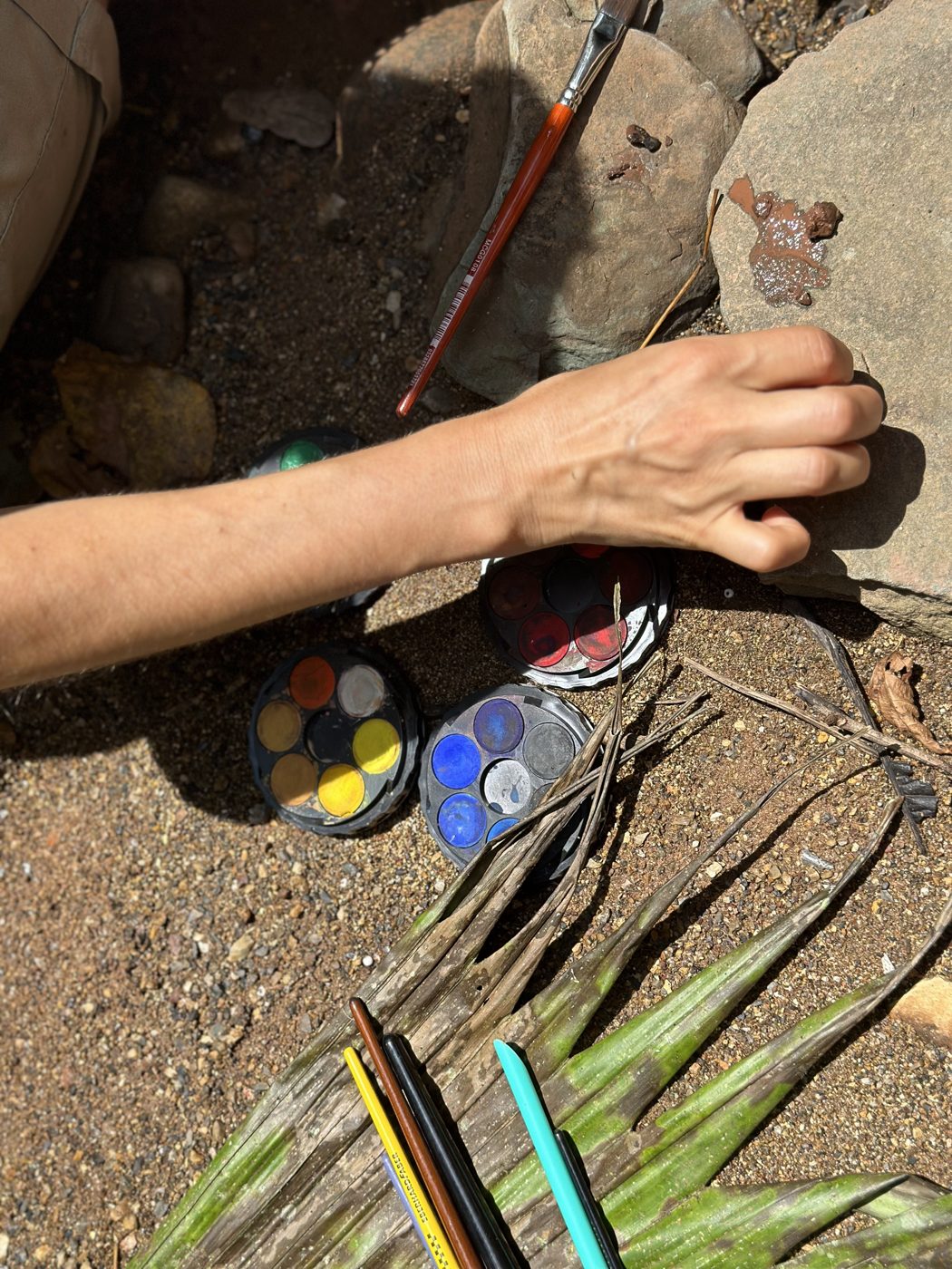 A hand painting with watercolors on a rock in the forest, with brushes, pencils and palm leaves nearby