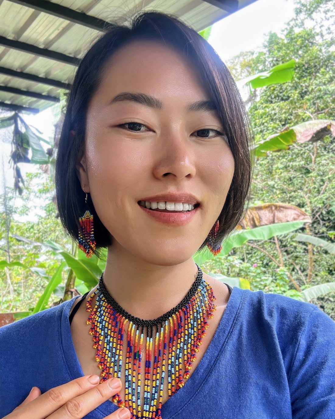 Min smiling on the porch in the jungle, wearing a colorful beaded necklace and earrings, one hand on her heart