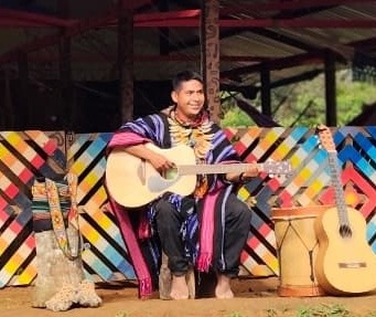 Taita Leonardo Jacanamejoy playing guitar in front of the colorful maloca wall